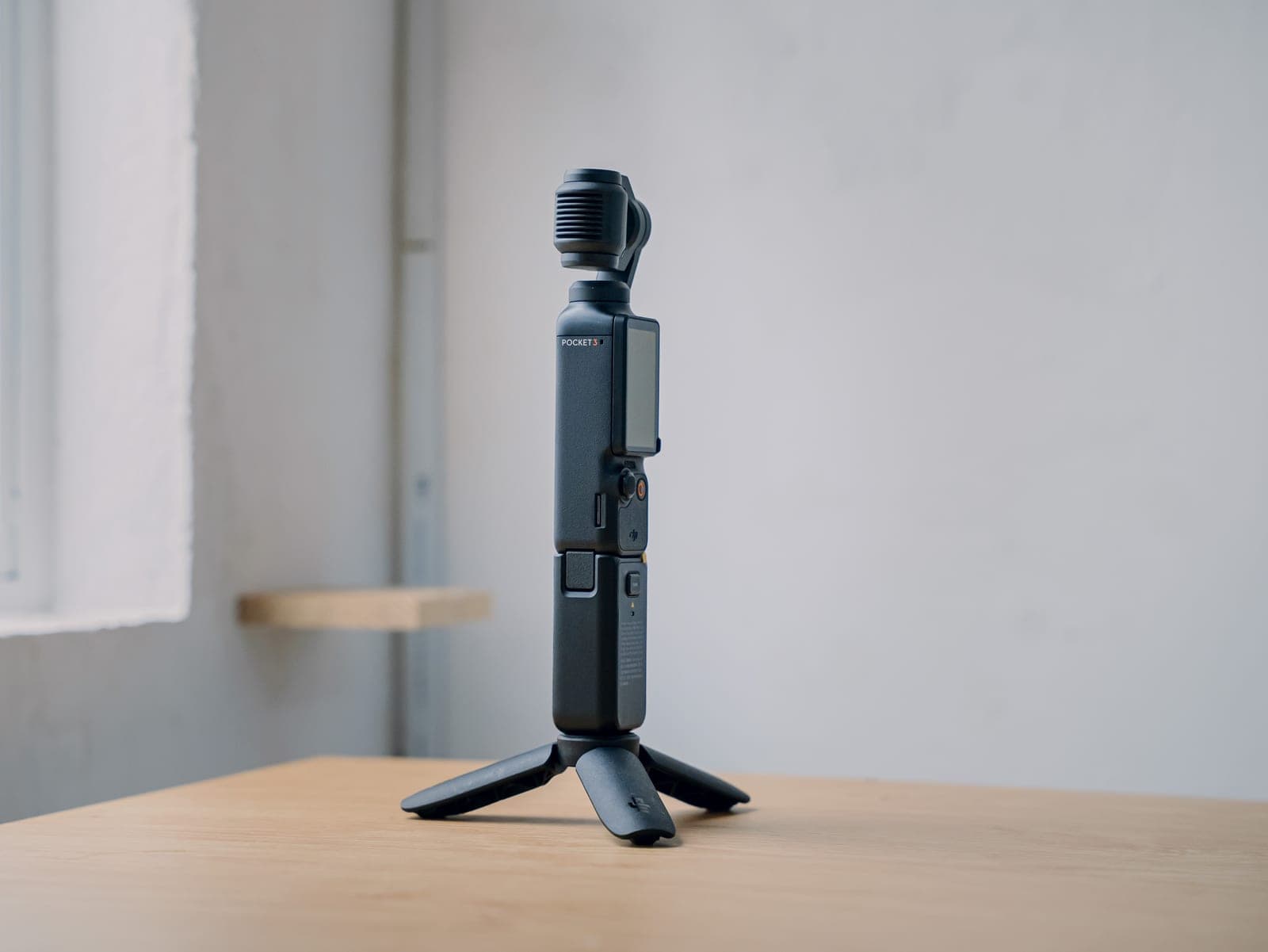 DJI Osmo Pocket 3 gắn trên chân đế mini, đặt trên mặt bàn gỗ cạnh cửa sổ