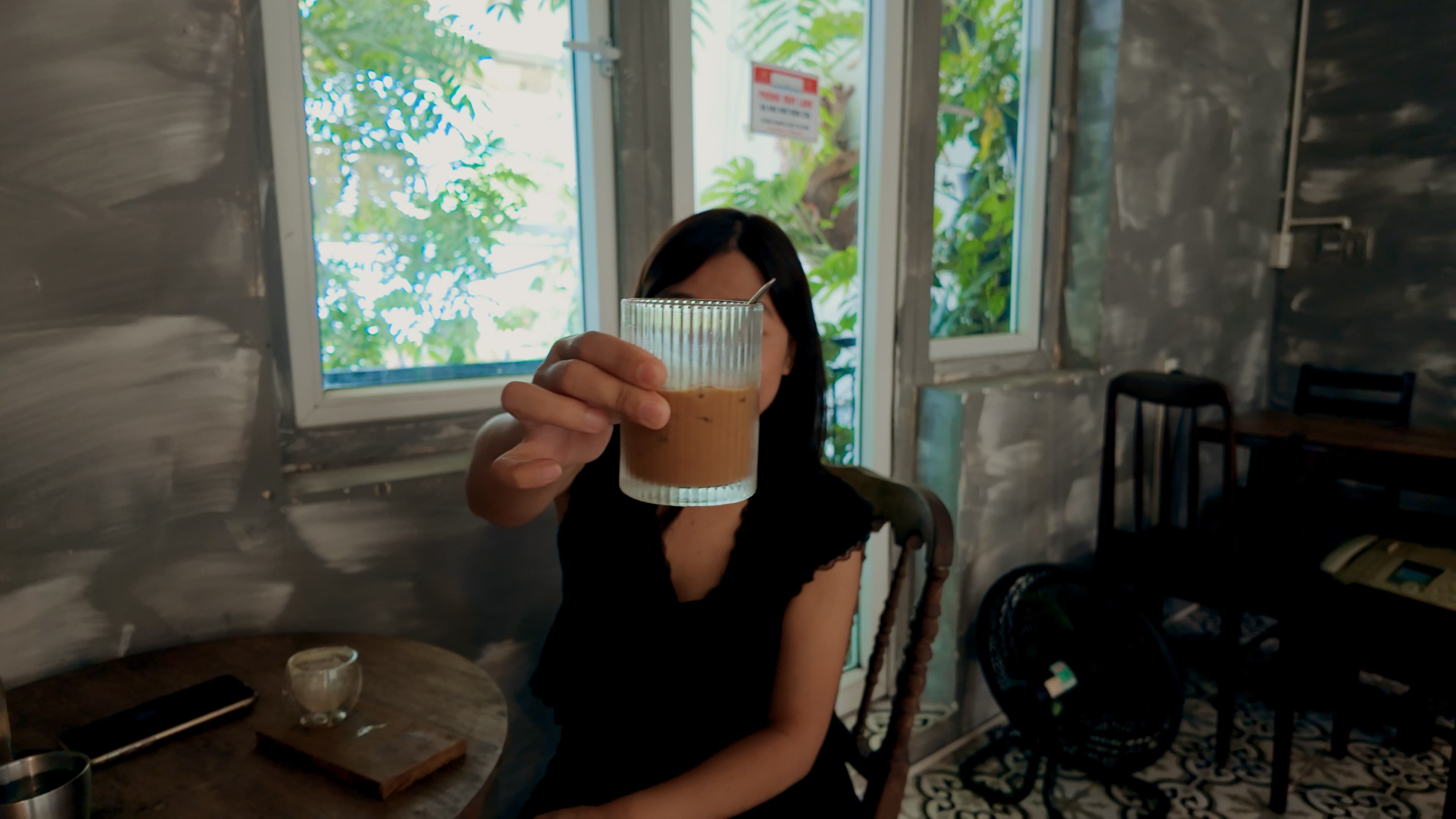 Xuân giơ ly cà phê về phía máy ảnh khi ngồi bên bàn gỗ trong quán, ánh sáng từ cửa sổ phía sau