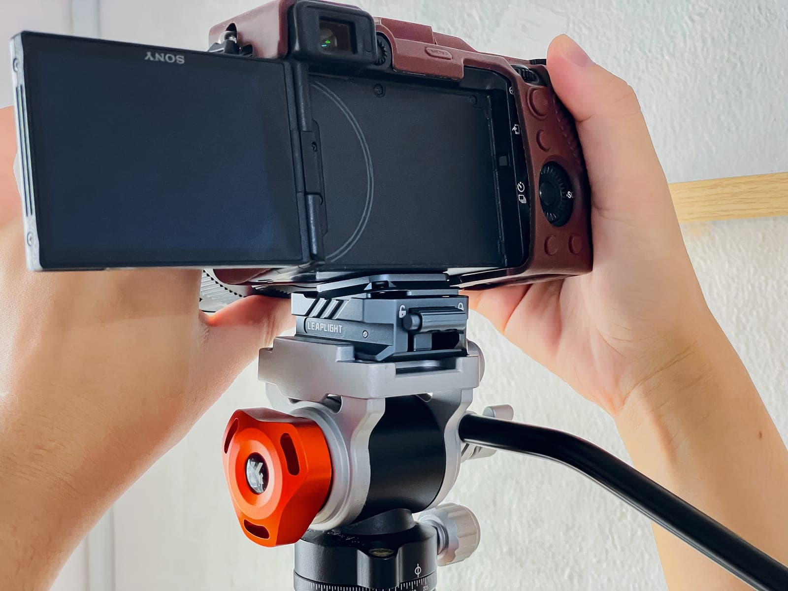 Hai tay gắn máy ảnh Sony lên mâm quick release của đầu tripod, màn hình máy đang lật ra