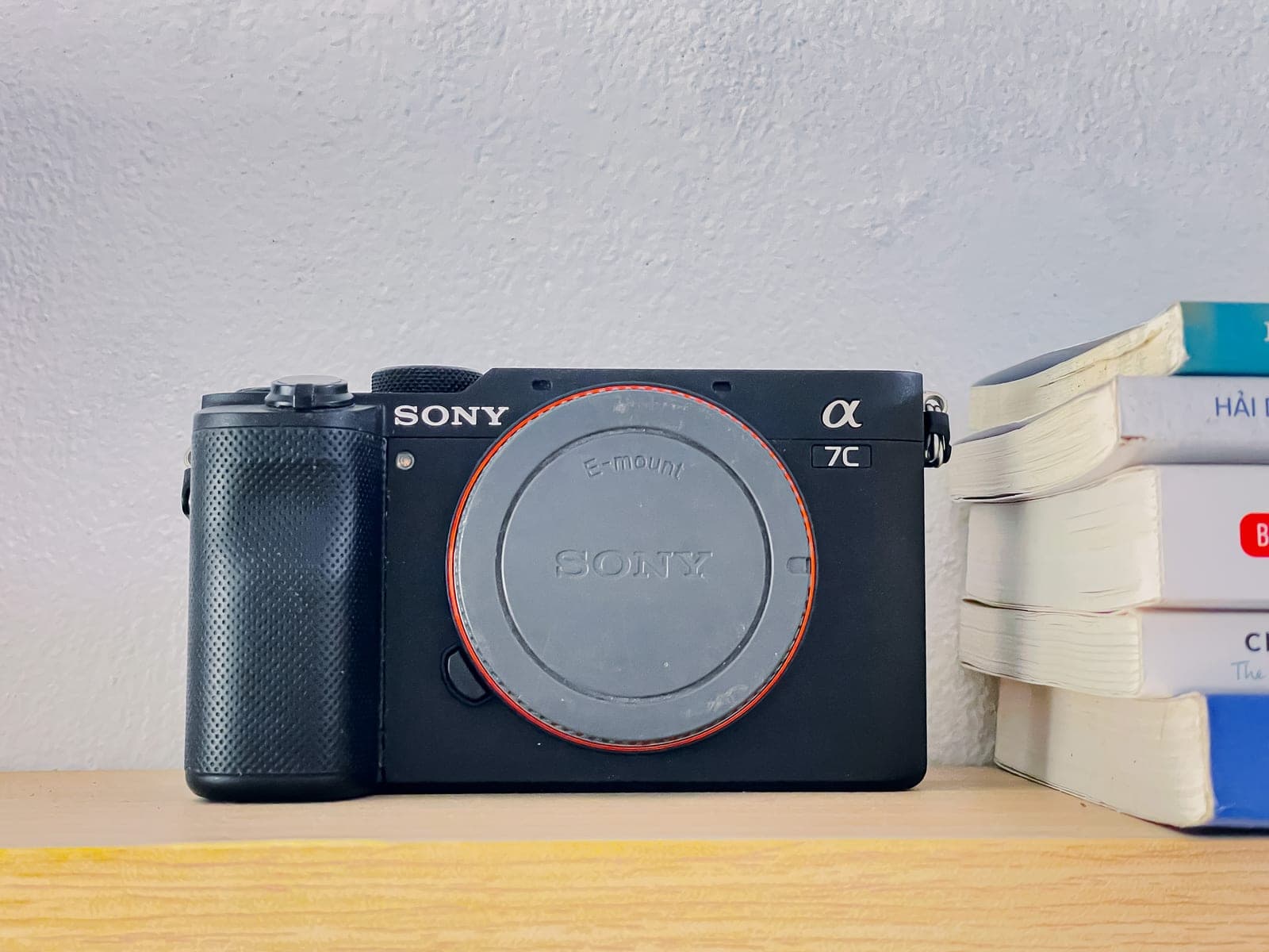 Thân máy ảnh Sony A7C đặt trên kệ gỗ sáng, bên cạnh một chồng sách, trên nền tường trắng